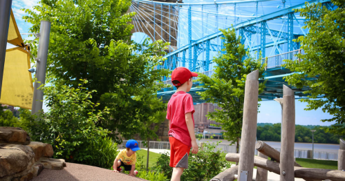 Two children play in a park with greenery, while a blue bridge spans over a river in the background.