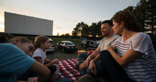 A family enjoys a picnic at a drive-in movie, sitting on a blanket with a screen and cars in the background.