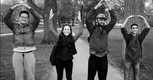 Four people stand on a path, forming the letter "O" with their arms, smiling in a park setting.