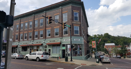 Historic brick building with shops on the ground floor, traffic lights, and a tree-lined street in a small town.