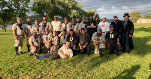 A group of vintage baseball players in period uniforms poses together on a grassy field, with trees in the background.