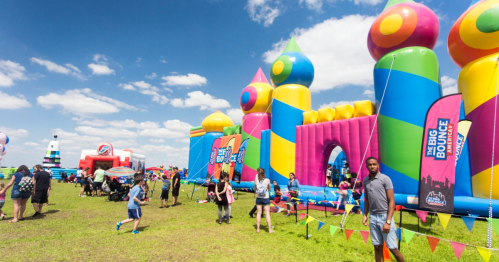 A colorful inflatable bounce castle with people enjoying a sunny day at a festival, surrounded by flags and other attractions.
