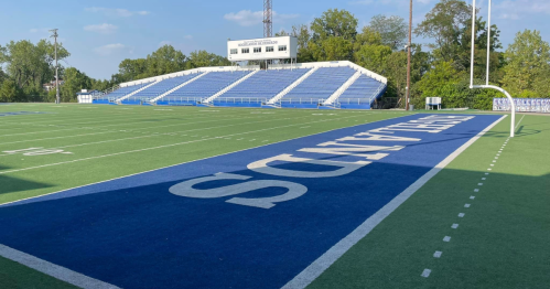 A football field with blue and white seating, featuring a goalpost and the word "STANDS" on the turf.