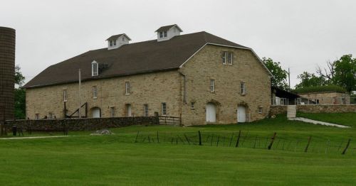 A large stone barn with a sloped roof, surrounded by green grass and a cloudy sky.