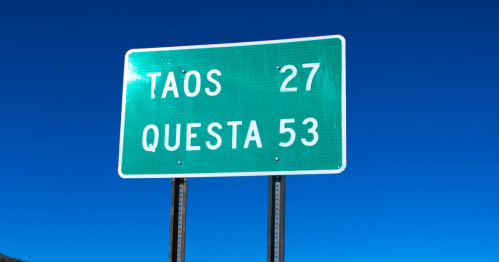 Green road sign indicating distances: Taos 27 miles, Questa 53 miles, against a clear blue sky.