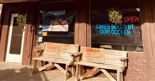 A storefront with an "OPEN" sign, featuring wooden benches outside and flower baskets, displaying an American flag.