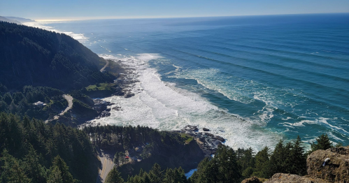 A scenic coastal view with waves crashing on the shore, surrounded by lush green forests and a clear blue sky.