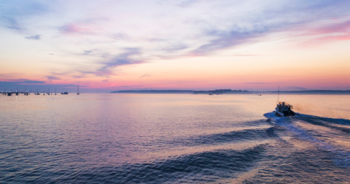 A boat glides across calm waters at sunset, with a colorful sky and distant boats in the background.