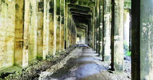 A long, empty corridor with tall, weathered pillars and a gravel path, surrounded by greenery.