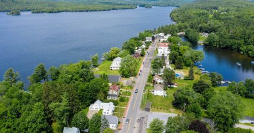 Aerial view of a lakeside town with green trees, houses, and a road along the water's edge.
