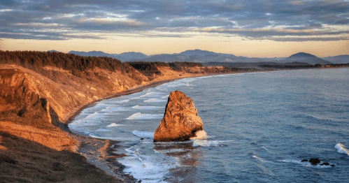 A scenic coastal view at sunset, featuring waves crashing against a rocky outcrop and rolling hills in the background.