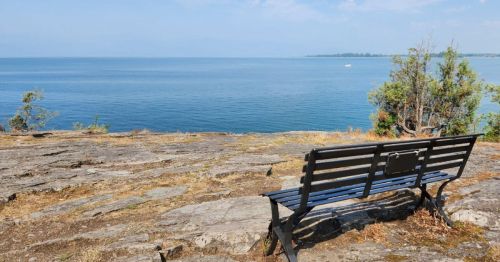 A solitary bench on a rocky shore overlooking a calm blue lake under a clear sky.
