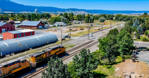 Aerial view of a rural area with train tracks, a yellow train, and buildings surrounded by trees and mountains in the background.