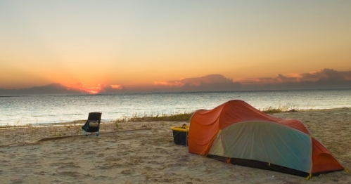 A tent and a chair on a sandy beach at sunset, with calm waters and colorful clouds in the sky.