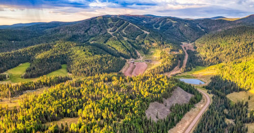 Aerial view of lush green mountains with a winding road, a small pond, and ski slopes in the distance under a blue sky.