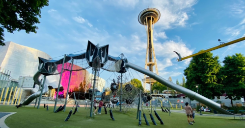 A playground structure with spider-like design in a park, with the Space Needle in the background under a blue sky.
