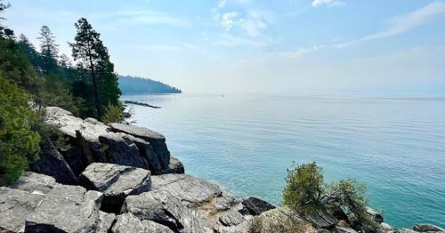 A serene lakeside view with rocky shores, trees in the background, and calm water under a clear sky.