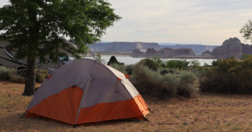 A tent with orange accents set up in a desert landscape, with mountains and a lake in the background.
