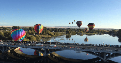 Colorful hot air balloons float over a river, with a crowd watching from a bridge below on a clear day.