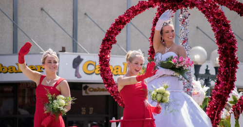 Three women in red dresses and gloves wave from a decorated float, with one in a white gown holding flowers.