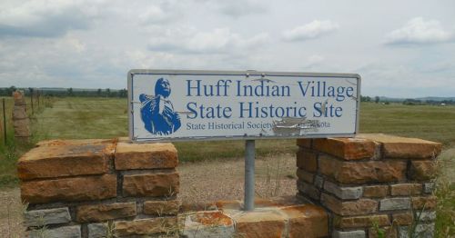 Sign for Huff Indian Village State Historic Site, featuring a depiction of a Native American figure, with a grassy field in the background.