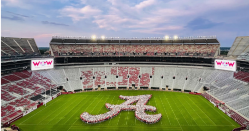 Aerial view of a stadium with a large group forming the letter "A" on the field, under a colorful sky.