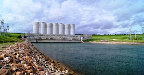 A large industrial facility with silos beside a calm body of water, surrounded by green hills and power lines.