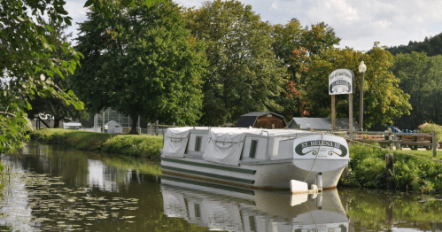 A boat named "St. Helena III" docked along a calm canal, surrounded by trees and a sign for a nearby establishment.