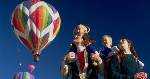 A family enjoys a hot air balloon festival, with colorful balloons in the sky and children on their shoulders.