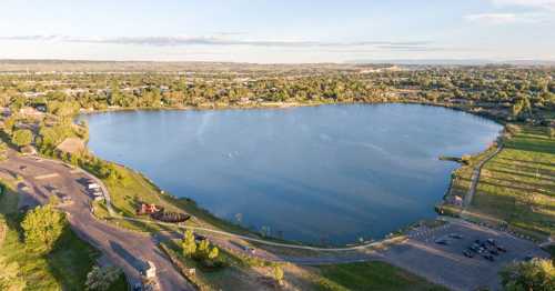 Aerial view of a serene lake surrounded by greenery and a parking area, with a clear sky in the background.