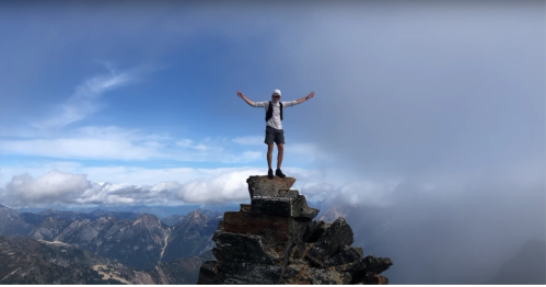 A person stands triumphantly on a rocky mountain peak, arms outstretched, with clouds and mountains in the background.