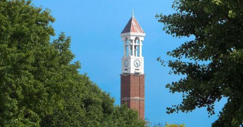 A clock tower with a peaked roof rises above green trees against a clear blue sky.