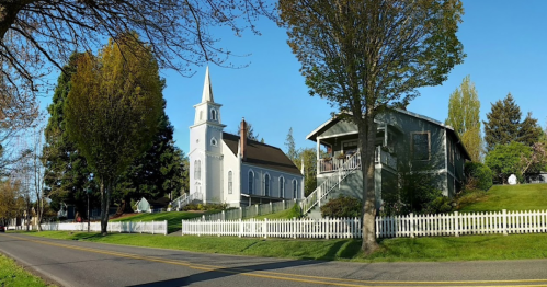 A scenic view of a church and a house beside a white picket fence, surrounded by trees on a sunny day.