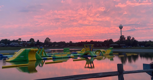 Colorful sunset over a lake with inflatable water park equipment and a water tower in the background.