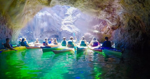 Group of kayakers inside a cave, surrounded by shimmering water and illuminated by sunlight streaming through the opening.
