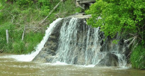A small waterfall cascading over rocks into a brown river, surrounded by lush green foliage.
