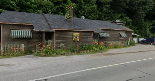 A rustic wooden building with green and white striped awnings, surrounded by greenery, along a roadside.