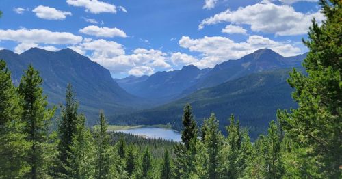 A scenic view of mountains and a lake surrounded by lush green trees under a blue sky with fluffy clouds.