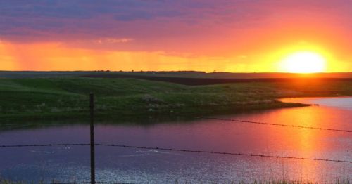 A vibrant sunset over a calm lake, with rolling hills and a barbed wire fence in the foreground.