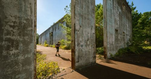 A person walks through the ruins of a concrete structure, surrounded by overgrown vegetation and trees.
