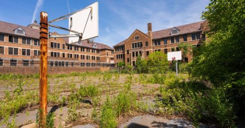 Abandoned basketball court overgrown with weeds, surrounded by a dilapidated brick building.