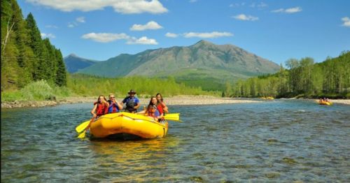 A group of people in a yellow raft on a river, surrounded by green trees and mountains under a blue sky.