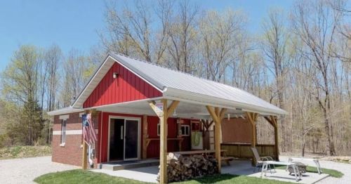 A red and white cabin with a metal roof, surrounded by trees and a gravel path, featuring a porch and outdoor seating.