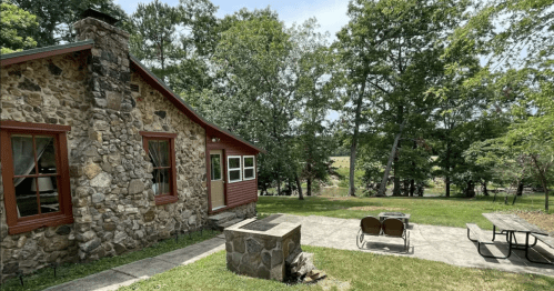 A rustic stone cabin surrounded by trees, with a patio and fire pit in a serene outdoor setting.