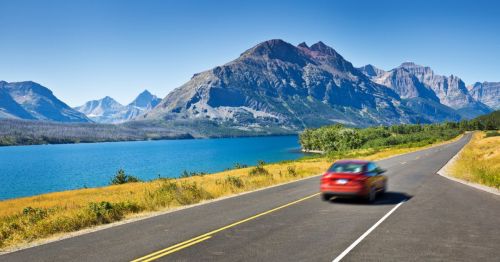 A red car drives along a scenic road beside a lake, with mountains in the background under a clear blue sky.