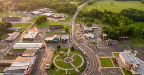 Aerial view of a small town with a roundabout, shops, and green fields in the background.