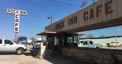 Exterior of Rock Inn Cafe with a sign, parking lot, and nearby vehicles on a clear day.