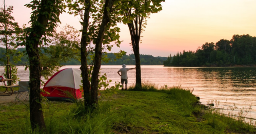 A person stands by a lake at sunset, with a tent and trees nearby, reflecting a peaceful camping scene.