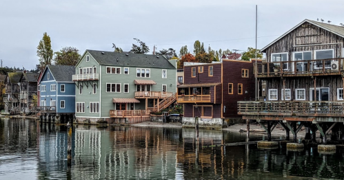 Colorful waterfront houses with wooden decks reflect in calm water under a cloudy sky.