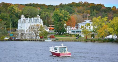 A small boat navigates a river near a scenic waterfront with colorful autumn trees and historic buildings.